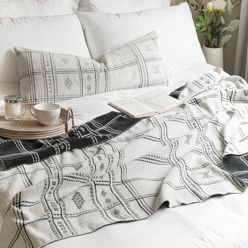 Puede incluir: Una cama blanca con una manta de tiro de patr&oacute;n blanco y negro. La manta tiene un dise&ntilde;o geom&eacute;trico. Hay dos almohadas blancas con fundas de almohada de patr&oacute;n blanco y negro en la cama. Un libro est&aacute; acostado sobre la manta.