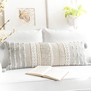 May include: A white couch with two white pillows and a long, gray and white patterned lumbar pillow. A book is lying open on the couch.