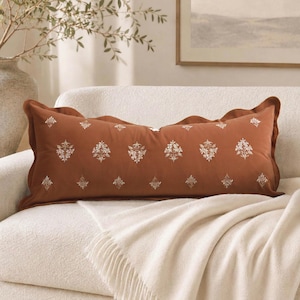 May include: A rectangular lumbar pillow in a burnt orange hue featuring a scalloped edge and a repeating pattern of small, white embroidered floral motifs across the front surface. The pillow has a soft, smooth texture and a finished border.
