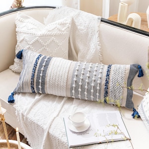 May include: A white couch with a white throw blanket and two pillows. One pillow is white with a textured design and the other is a long, rectangular pillow with a blue and white striped pattern. The striped pillow has a tassel on each end.