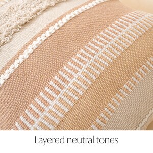May include: Close-up of a decorative pillow with layered neutral tones. The pillow features a textured design with alternating stripes of beige, cream, and peach. White stitching and fringe add detail to the woven fabric.