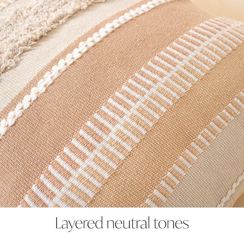 May include: Close-up of a decorative pillow with layered neutral tones. The pillow features a textured design with alternating stripes of beige, cream, and peach. White stitching and fringe add detail to the woven fabric.