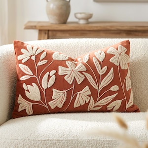 May include: A rectangular throw pillow in a deep terracotta orange color featuring raised, textured cream-colored embroidery of various floral and botanical patterns, including large blossoms, buds, and leaves across the front surface.