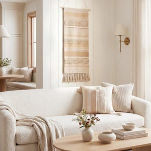 May include: A neutral-toned living room scene featuring a white sofa with decorative pillows and a throw blanket. A beige wall hanging with tassels is displayed above the sofa. A wooden coffee table holds a vase with flowers and decorative bowls.