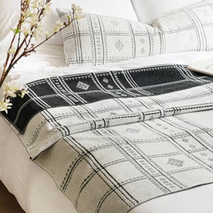 Puede incluir: Una manta de tiro con estampado en blanco y negro con un dise&ntilde;o geom&eacute;trico. La manta est&aacute; doblada por la mitad y colocada sobre una cama blanca con dos almohadas.