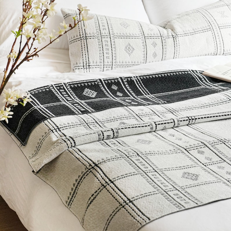 Puede incluir: Una manta de tiro con estampado en blanco y negro con un dise&ntilde;o geom&eacute;trico. La manta est&aacute; doblada por la mitad y colocada sobre una cama blanca con dos almohadas.