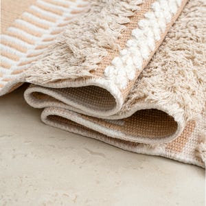 May include: Rolled-up beige and white patterned rug. The rug features a geometric design with a fringe border. The color palette includes beige, cream, and white, creating a neutral and textured appearance. The rug is made of a woven material.