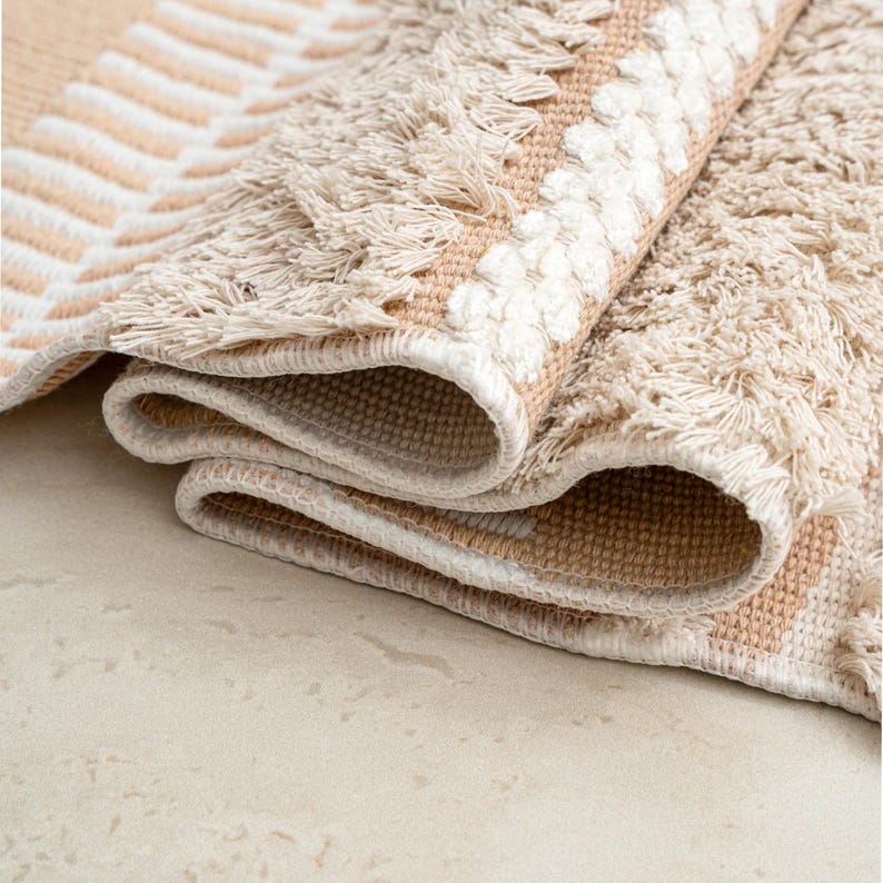 May include: Rolled-up beige and white patterned rug. The rug features a geometric design with a fringe border. The color palette includes beige, cream, and white, creating a neutral and textured appearance. The rug is made of a woven material.