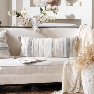 May include: A white couch with a gray and white patterned lumbar pillow. The couch is decorated with a white throw blanket and a bouquet of white flowers.