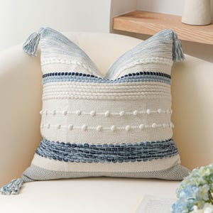 May include: A square throw pillow featuring a textured cream-colored base with horizontal stripes in shades of light blue, navy, and gray. The design includes woven patterns, raised white dots, and light blue tassels at the corners.