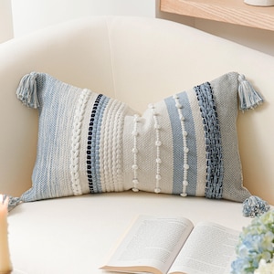 May include: A rectangular decorative pillow with a textured design in shades of blue, white, and gray. The pillow features stripes, braided details, and small tufts. Tassels adorn each corner. The pillow is on a white chair.