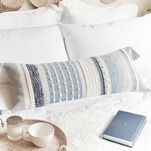 May include: A long, rectangular throw pillow with a blue, white, and beige striped pattern. The pillow has a textured surface with small white pom-poms and fringe details.