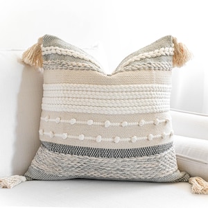 May include: A decorative throw pillow with a woven design in shades of gray, beige, and white. The pillow has a fringe trim and features a textured pattern with small white pom-poms.