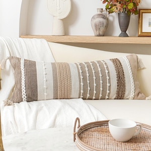 May include: A rectangular lumbar pillow featuring a textured, woven design in shades of cream, beige, and brown. The pillow displays vertical stripes with varying patterns, including braided accents and small tufted dots, finished with tassels.