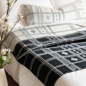Puede incluir: Manta de tiro de patr&oacute;n blanco y negro con un dise&ntilde;o geom&eacute;trico. La manta est&aacute; doblada en una cama blanca con dos almohadas blancas con fundas de almohada de patr&oacute;n blanco y negro.