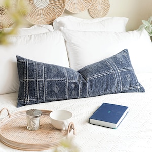 May include: A navy blue lumbar pillow with a white geometric pattern rests on a white bed. A woven tray holds a candle, a white teacup, and a blue book. Woven wall decor is visible in the background.