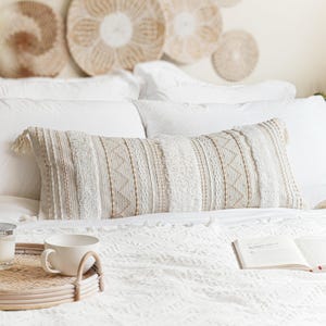 May include: A white and beige patterned throw pillow with fringe on the ends. The pillow is on a white bed with a white blanket and a book on top of the blanket.