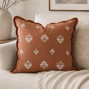 May include: A square terracotta-colored throw pillow featuring a repeating pattern of small, white embroidered floral sprigs. The pillow has a ruffled flange edge and a soft, matte fabric texture, presenting a consistent design across the front.