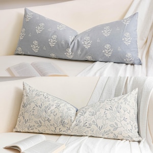May include: Two decorative lumbar pillows with floral patterns rest on a white sofa. One pillow is light blue with white floral designs. The other pillow is off-white with blue floral and leaf patterns. Open books are visible.