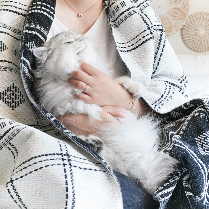 Puede incluir: Una persona con una manta de patr&oacute;n blanco y negro sostiene un gato blanco y esponjoso en su regazo.