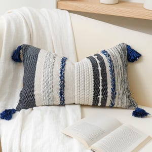 May include: A rectangular throw pillow featuring a textured pattern of cream, black, and navy blue stripes. The design includes braided navy blue accents, raised dot textures, and a solid navy blue tassel at each of the four corners.