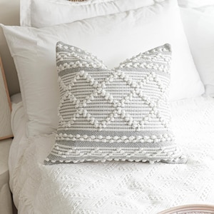 May include: A gray and white patterned throw pillow with a textured surface. The pillow has a diamond pattern with white pom-poms.