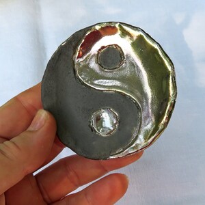 May include: A Yin and Yang symbol, a ceramic piece with a dark gray base and a shiny silver-colored design. The symbol is round with a raised edge. The Yin and Yang symbol is a representation of balance and harmony.