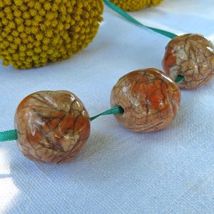 May include: Three round, glossy ceramic beads with a crackled texture in shades of tan, brown, and orange, strung on a thin green ribbon. The beads feature a distinct web-like pattern across their surfaces, showcasing a smooth, reflective finish.