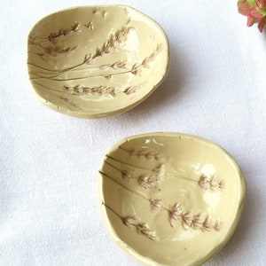 May include: Two small, irregular, cream-colored ceramic bowls featuring an embossed lavender sprig design in soft brown tones. The bowls have a glossy finish and organic, hand-formed edges, showcasing a delicate botanical motif on the interior surface.
