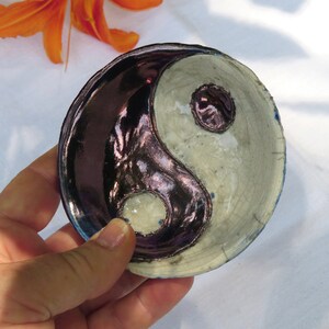 May include: A small, round ceramic dish with a yin-yang design. The dish is divided into two sections: one is a dark, glossy purple, and the other is a light, crackled cream color. The dish is held in a hand.