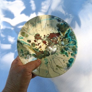 May include: A round ceramic bowl featuring a crackle glaze finish in cream, accented with vibrant splashes of turquoise, deep green, and rust-red. The surface displays a network of dark, thin lines radiating across the interior, creating a textured appearance with a glossy, reflective sheen throughout the piece.