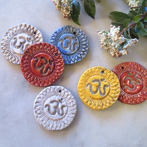 May include: Six circular ceramic pendants featuring a raised Om symbol in the center and a sunburst pattern around the edge. The pendants are glazed in white, blue, red, and yellow, each with a small hole at the top for hanging or stringing.