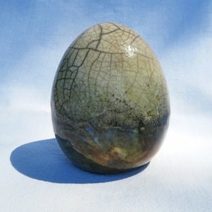 Pode incluir: Uma escultura de cerâmica em forma de ovo com acabamento brilhante e um padrão de esmalte craquelado. A parte superior é de cor creme pálido, transitando para uma mistura escura e manchada de tons verde-escuro, castanho e azul na base.