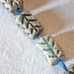 Può includere: Primo piano di una collana di perline in ceramica fatta a mano. Le perline sono rettangolari con disegni di foglie nei toni del blu e del marrone. Le perline sono infilate su un nastro di raso blu. Lo sfondo è un tessuto bianco strutturato.