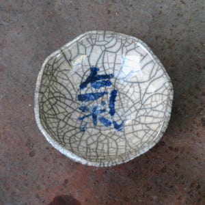 May include: A small, off-white ceramic dish with a crackle glaze pattern. The dish has a slightly irregular shape and features blue Japanese characters in the center. The dish is likely used for serving small portions of food.