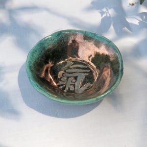 May include: A small, handmade ceramic bowl with a teal and bronze glaze. The bowl features an embossed Japanese character in the center. The rim has an uneven, organic shape. The bowl is set against a white background.