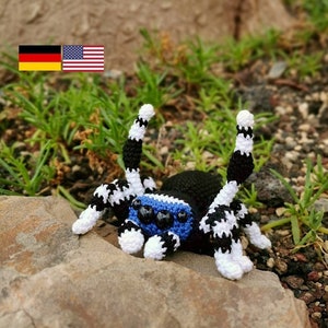 Könnte beinhalten: Ein gehäkeltes Sp Spielzeug mit einem schwarzen Körper, blauen Augen und schwarz-weiß gestreiften Beinen. Die Spinne sitzt auf einem Felsen, mit grünem Gras und einem Baumstamm im Hintergrund.