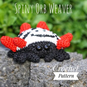 Könnte beinhalten: Ein Häkelmuster für eine stachelige Kugelnetzspinne. Die Spinne ist schwarz, weiß und rot mit großen schwarzen Augen. Die Spinne sitzt auf einem Stein.