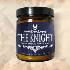 May include: A glass jar candle with a black lid. The label is navy blue with white text that reads "THE KNIGHT" and "CAVERN WATER, MINERALS & MOSS." The candle is 8 oz and made with hand-poured soy blend wax.