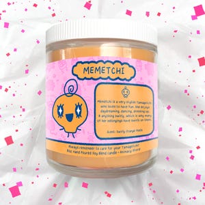 May include: A glass jar candle with a white lid and orange wax. The label features a cartoon character and the text "MEMETCHI". The scent is "Swirly Orange Peels". The candle is 8 oz.
