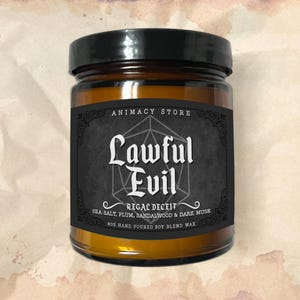May include: A brown glass candle jar with a black label. The label has a black and white image of a Dungeons and Dragons die with the text "Lawful Evil" and "Regal Deceit" written on it. The label also lists the scent notes: "Sea Salt, Plum, Sandalwood & Dark Musk". The bottom of the label reads "8oz Hand Poured Soy Blend Wax".