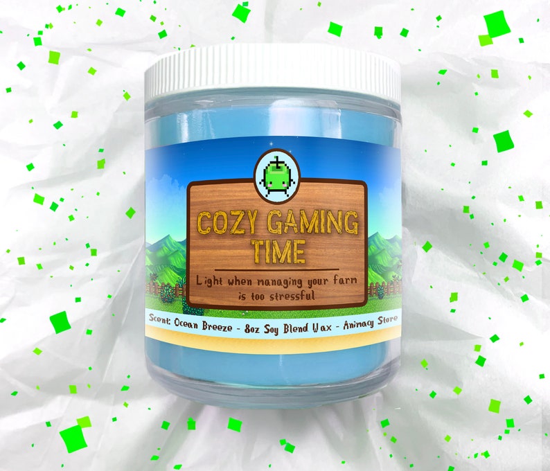 May include: A blue glass candle jar with a white lid. The label says "Cozy Gaming Time" with a pixelated green character. The text below says "Light when managing your farm is too stressful." The scent is "Ocean Breeze" and the candle is 8oz Soy Blend Wax.