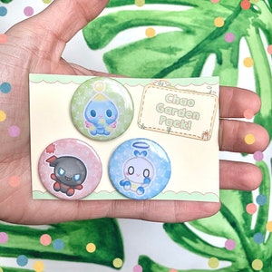 May include: Three round button pins featuring cartoon characters from the video game series Sonic the Hedgehog. The pins are on a white card with the text "Chao Garden Pack!" in a yellow banner.