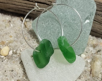 May include: A pair of silver hoop earrings with green sea glass accents. The earrings are displayed on a white sea glass stone.