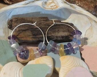 May include: A pair of silver hoop earrings with a colorful assortment of gemstone chips. The chips are shades of purple, blue, yellow, and green.