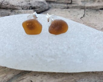 May include: A pair of earrings featuring two small, amber-colored sea glass pieces set in silver.
