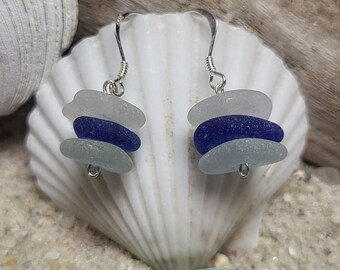 May include: A pair of silver dangle earrings with three stacked sea glass beads. The beads are white, blue, and white. The earrings are displayed on a white seashell.