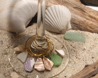 May include: A gold-colored wine glass charm with five different sea-themed charms hanging from it. The charms include a purple and white seashell, a pink seashell, a green piece of sea glass, and two white seashells.