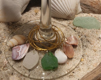 May include: A glass wine glass with a gold wire wrapped around the stem. The wire is decorated with seashells and sea glass. The glass is sitting on a bed of sand.