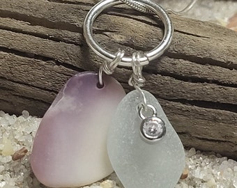 May include: A silver necklace featuring two sea glass pendants. One pendant is a soft lavender color, the other is a pale green with a small, clear gemstone. The pendants are suspended from a silver ring and chain.